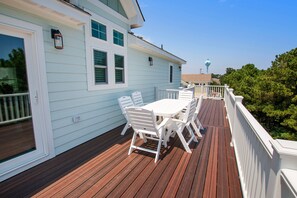 Une terrasse en bois avec une table et des chaises blanches, une balustrade blanche et un bâtiment bleu clair doté de fenêtres.