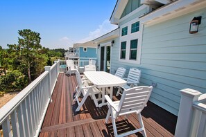 Une terrasse en bois avec des meubles d’extérieur blancs, une rambarde et un bâtiment bleu clair doté de fenêtres.