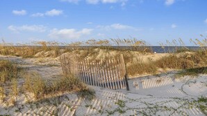 Une plage de sable avec des dunes, une clôture en bois et un ciel bleu azur.