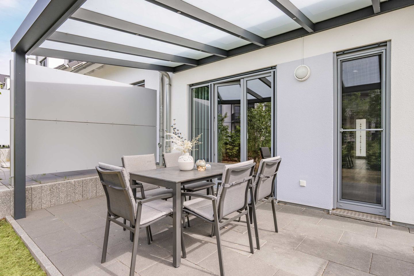 Eine Terrasse im Freien mit Tisch und Stühlen, einer Glastür und einem überdachten Bereich mit Metallgestell.