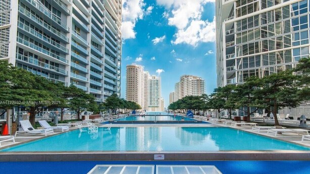 A swimming pool with lounge chairs and a glass table in the foreground, surrounded by tall buildings and trees.