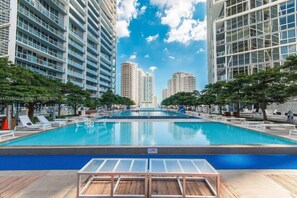 A swimming pool with lounge chairs and a glass table in the foreground, surrounded by tall buildings and trees.