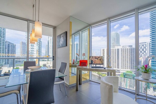 A modern office space with a glass table, chairs, a desk with a computer, and a large window offering a city view.