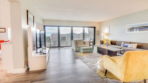 A modern living room with a sofa, armchair, coffee table, and a dining area with a TV and bicycle. - Top-floor Condo w/ Stunning Views, 2 Pools & More (New Smyrna Beach)