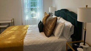 Een net opgemaakt bed met een gouden deken, een groen bekleden hoofdeinde en een nachtkastje met een lamp.