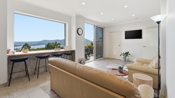 A modern living room with a large window, a sofa, a coffee table, and a TV mounted on the wall. - MONA Vista, overlooking Museum of Old and New Art (MONA) & the River Derwent. (Rosetta)