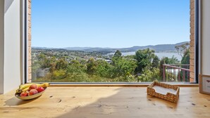 A room with a large window overlooking a scenic landscape, a wooden table with a bowl of fruit, and a woven basket. - MONA Vista, overlooking Museum of Old and New Art (MONA) & the River Derwent. (Rosetta)