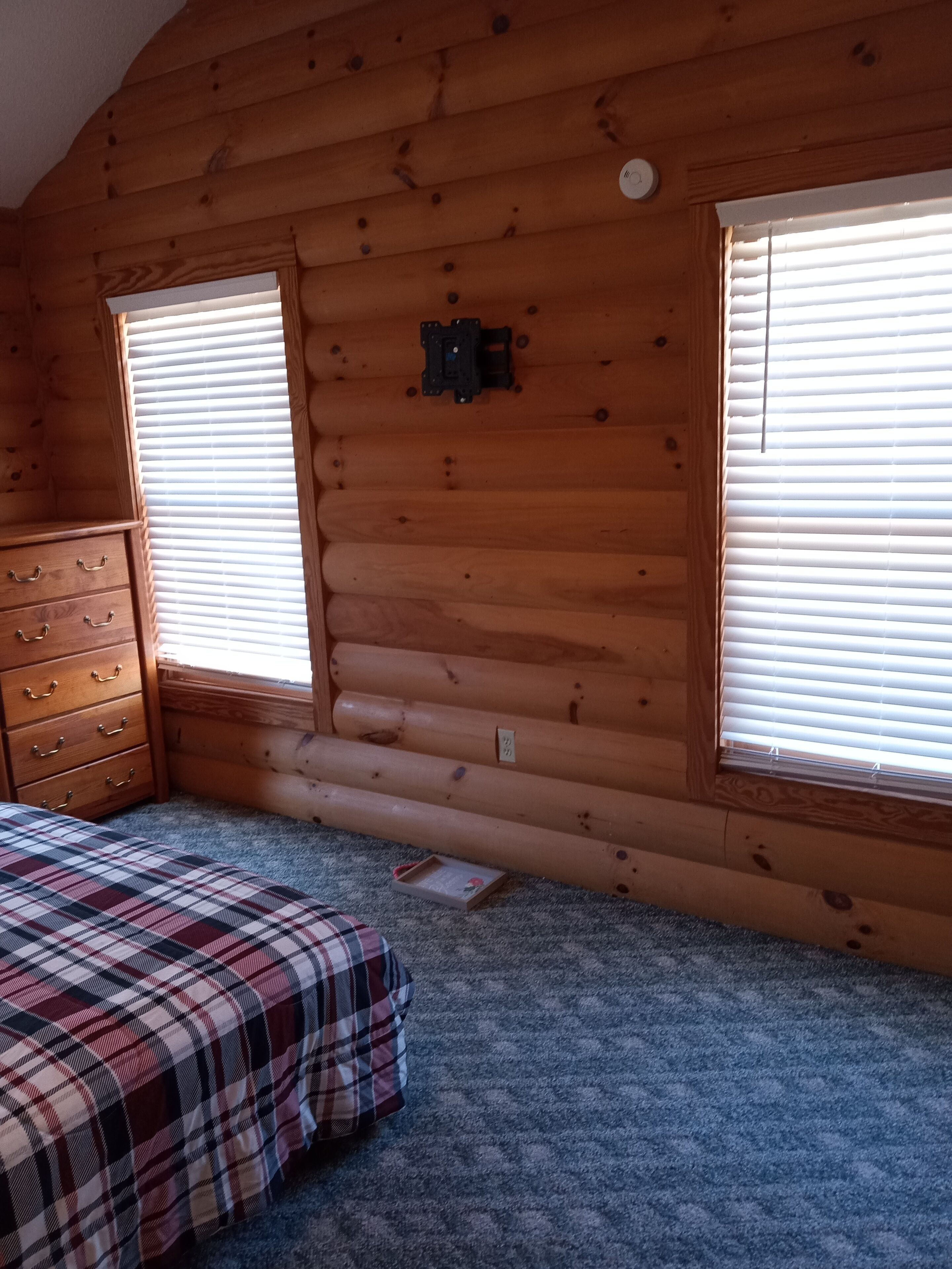 Een slaapkamer in een blokhut met een geruit sprei, een houten dressoir en twee ramen met jaloezieën.