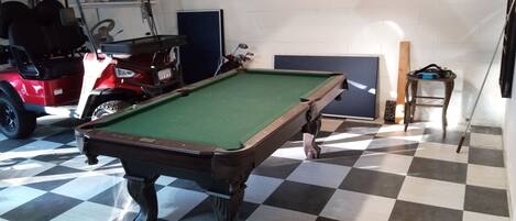 A pool table in a room with a checkered floor, a lamp, and a red vehicle in the background.