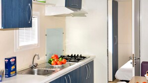 Une cuisine de taille réduite, équipée d’une cuisinière à gaz, d’un évier et de meubles de rangement bleus. Une fenêtre avec des stores et une porte donnant sur une autre pièce se trouvent également dans la cuisine.