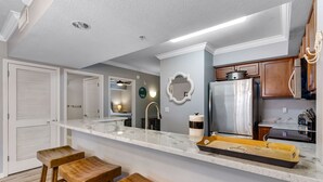 A modern kitchen with a marble countertop, wooden stools, a stainless steel refrigerator, and wooden cabinets. - ☀️ Pelican 307: Your VIP Beach Getaway Awaits | Lazy River & FREE Golf (Fort Walton Beach)