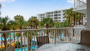 A balcony with a glass table, chairs, and a view of a swimming pool and palm trees. - ☀️ Pelican 307: Your VIP Beach Getaway Awaits | Lazy River & FREE Golf (Fort Walton Beach)