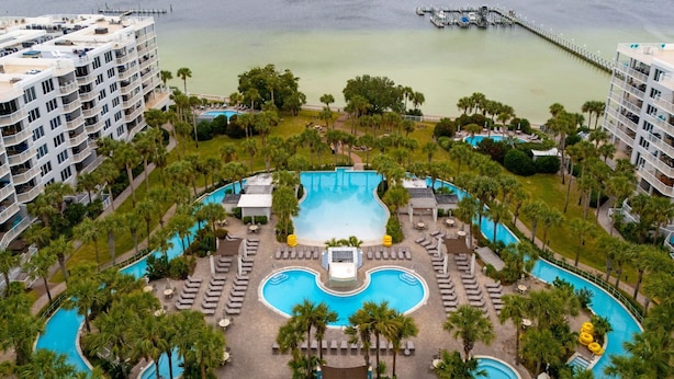 A resort with a central swimming pool surrounded by lounge areas and palm trees, with a body of water and a pier in the background. - ☀️ Pelican 307: Your VIP Beach Getaway Awaits | Lazy River & FREE Golf (Fort Walton Beach)