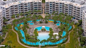 A high-rise hotel with a central swimming pool surrounded by palm trees and lounge areas. - ☀️ Pelican 307: Your VIP Beach Getaway Awaits | Lazy River & FREE Golf (Fort Walton Beach)