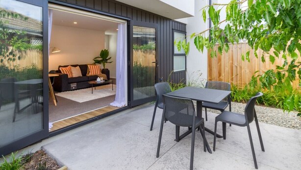 Un patio moderno con una puerta de vidrio que da a una sala de estar, una mesa con sillas y una valla de madera al fondo.