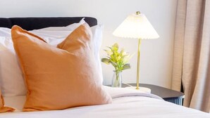 Una cama bien hecha con almohadas blancas y color melocotón, una toalla blanca enrollada y una lámpara de noche en una mesita.