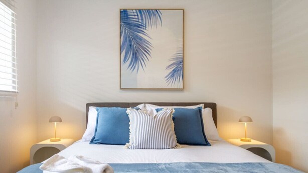Una cama bien tendida con ropa de cama azul y blanca, dos lámparas de noche y una obra de arte enmarcada en la pared.