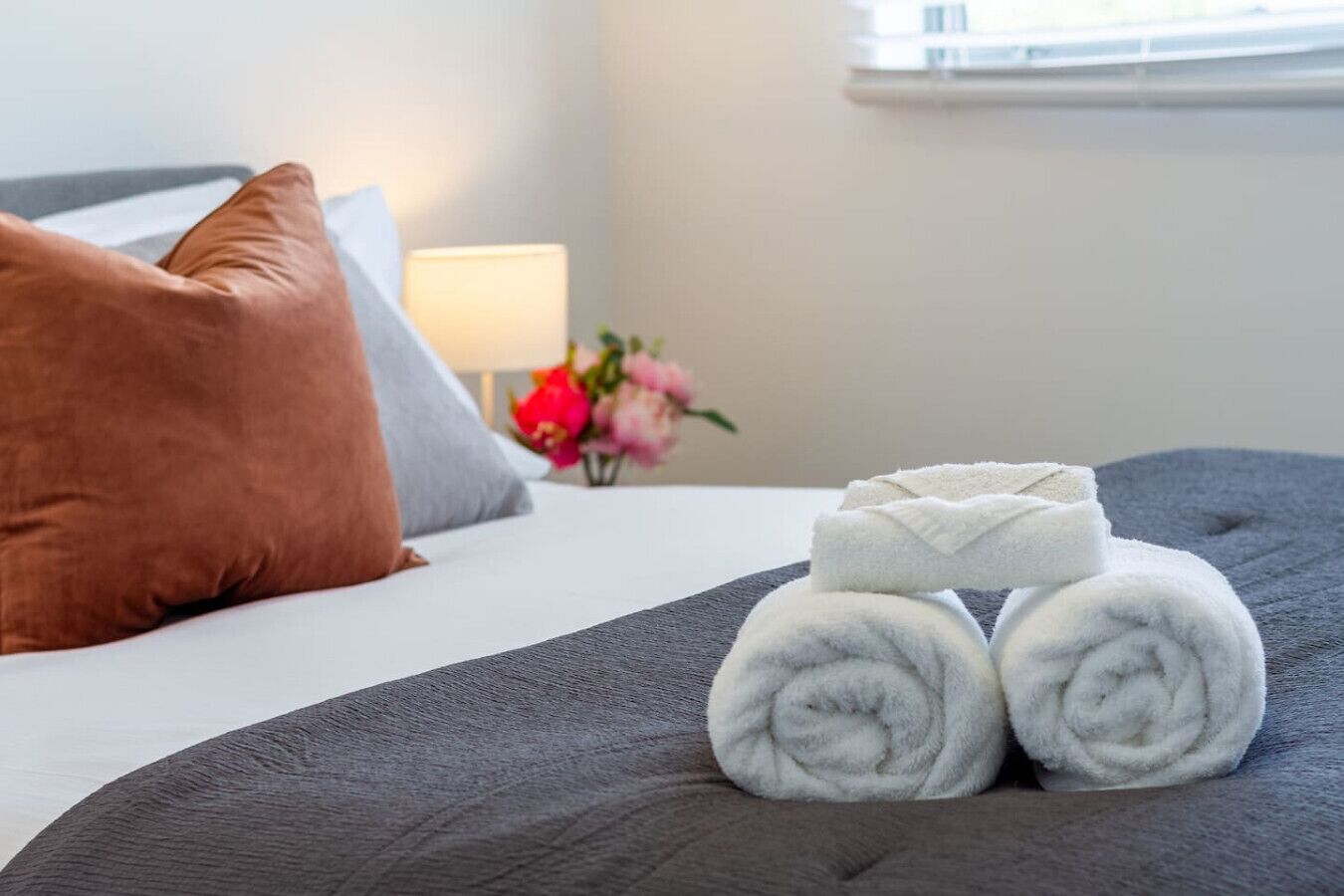 Una cama bien hecha con una manta gris, dos toallas blancas enrolladas y una almohada marrón.