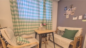 Ein gemütliches Zimmer mit einem karierten Vorhang, einem kleinen Tisch mit Blumen in einer Vase, einem Sofa mit einem leopardenmuster-Kissen und einer Wand, die mit verschiedenen Bildern und einem Thermostat verziert ist.