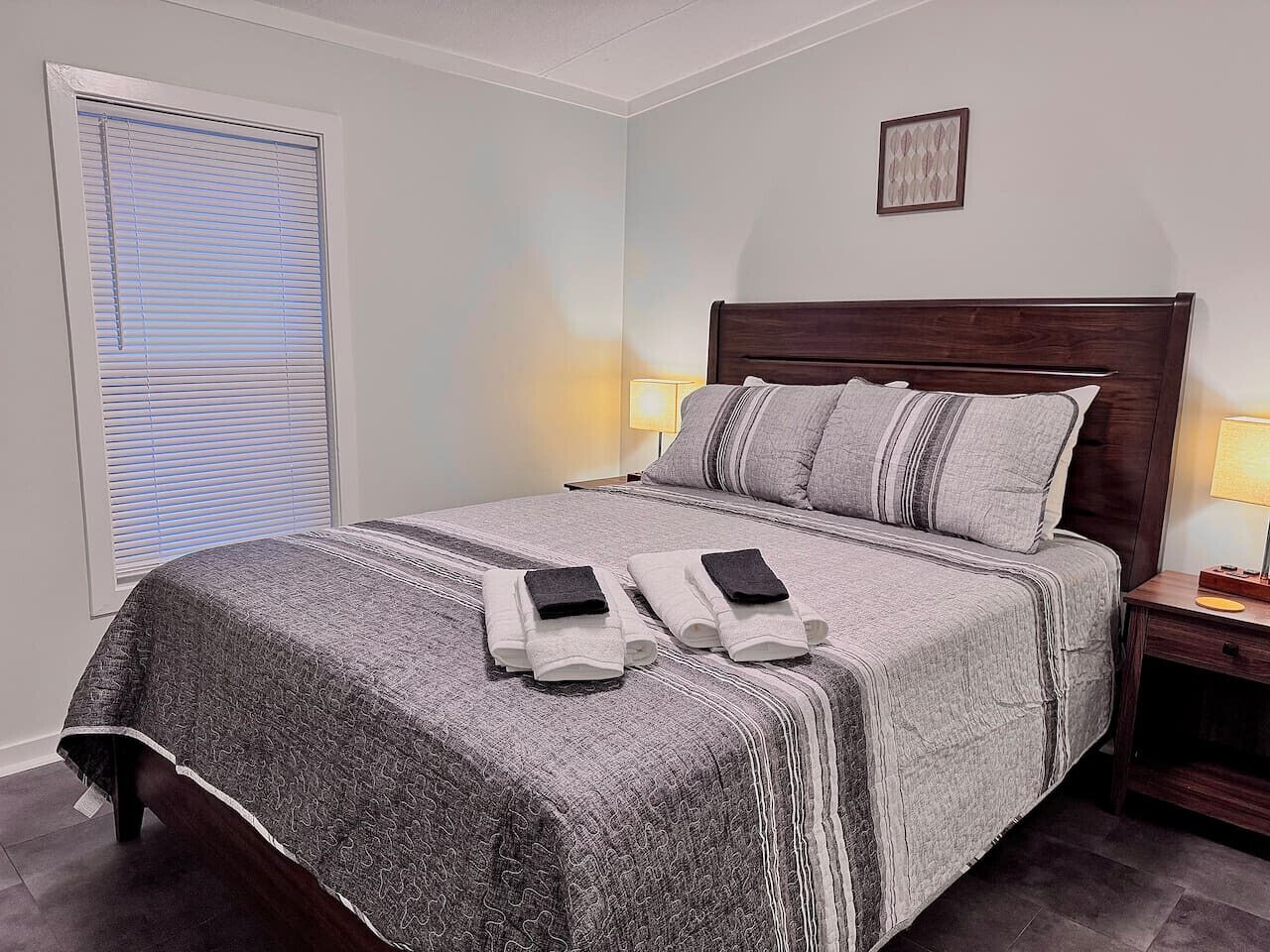 A neatly made bed with a patterned bedspread, two pillows, and two folded towels on the bed. There is a bedside table with a lamp and a framed picture on the wall above the bed.