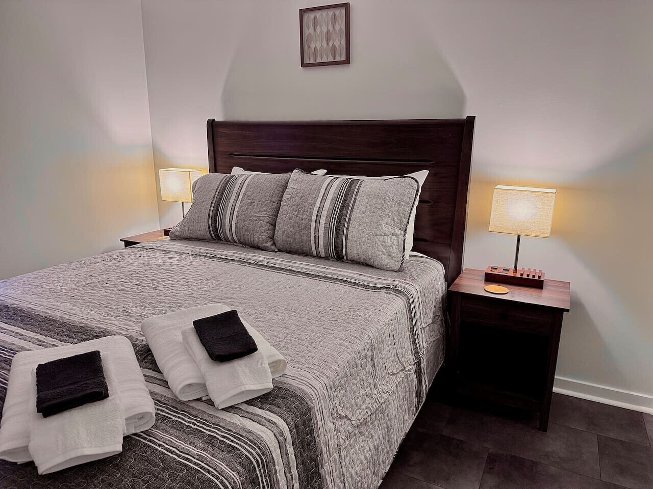 A neatly made bed with striped bedding and pillows, two folded towels, a wooden nightstand with a lamp, and a framed picture on the wall.