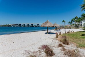 Una playa con sombrillas de paja, un cielo azul despejado y un puente al fondo.