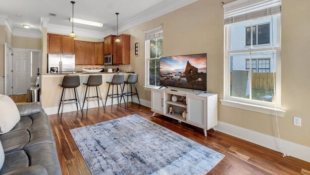 A modern kitchen with wooden cabinets, a central island with bar stools, a large flat-screen TV, and a view of the backyard through a large window. - Midtown Home, Pets Welcome! (Tallahassee)