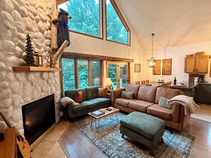 A spacious living room with a stone fireplace, a large window overlooking greenery, a cozy seating area, and a kitchen in the background.