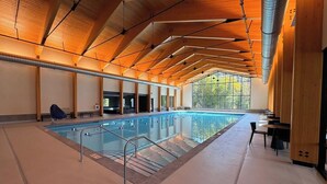 An indoor swimming pool with a high ceiling and wooden beams, surrounded by seating areas.