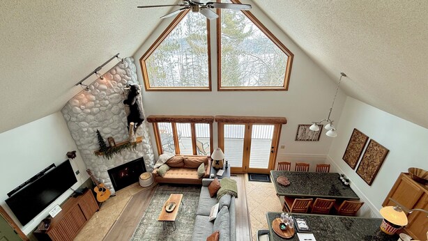 A spacious living area with a stone fireplace, a large window, a ceiling fan, and a dining area with a kitchen in the background.