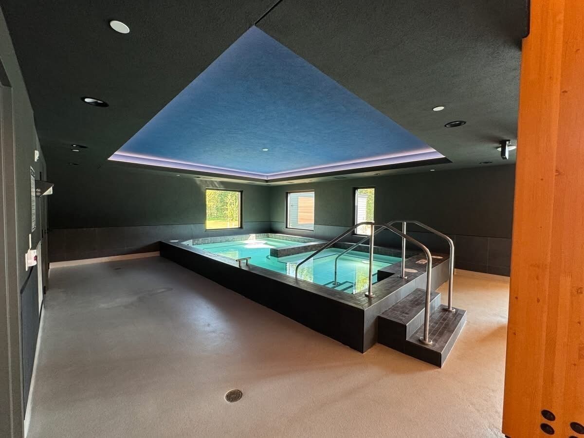 A modern indoor swimming pool with a skylight, surrounded by dark walls and a concrete floor.