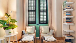 A room with two wicker chairs, a small table with a potted plant, a bookshelf, and a window with green shutters.