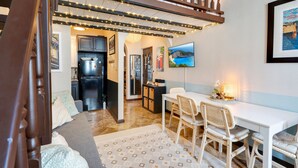 A cozy living area with a sofa, a dining table with chairs, a kitchen in the background, and a TV mounted on the wall.