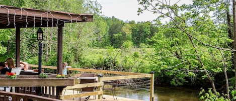A wooden deck with chairs and tables, overlooking a river and surrounded by greenery.
