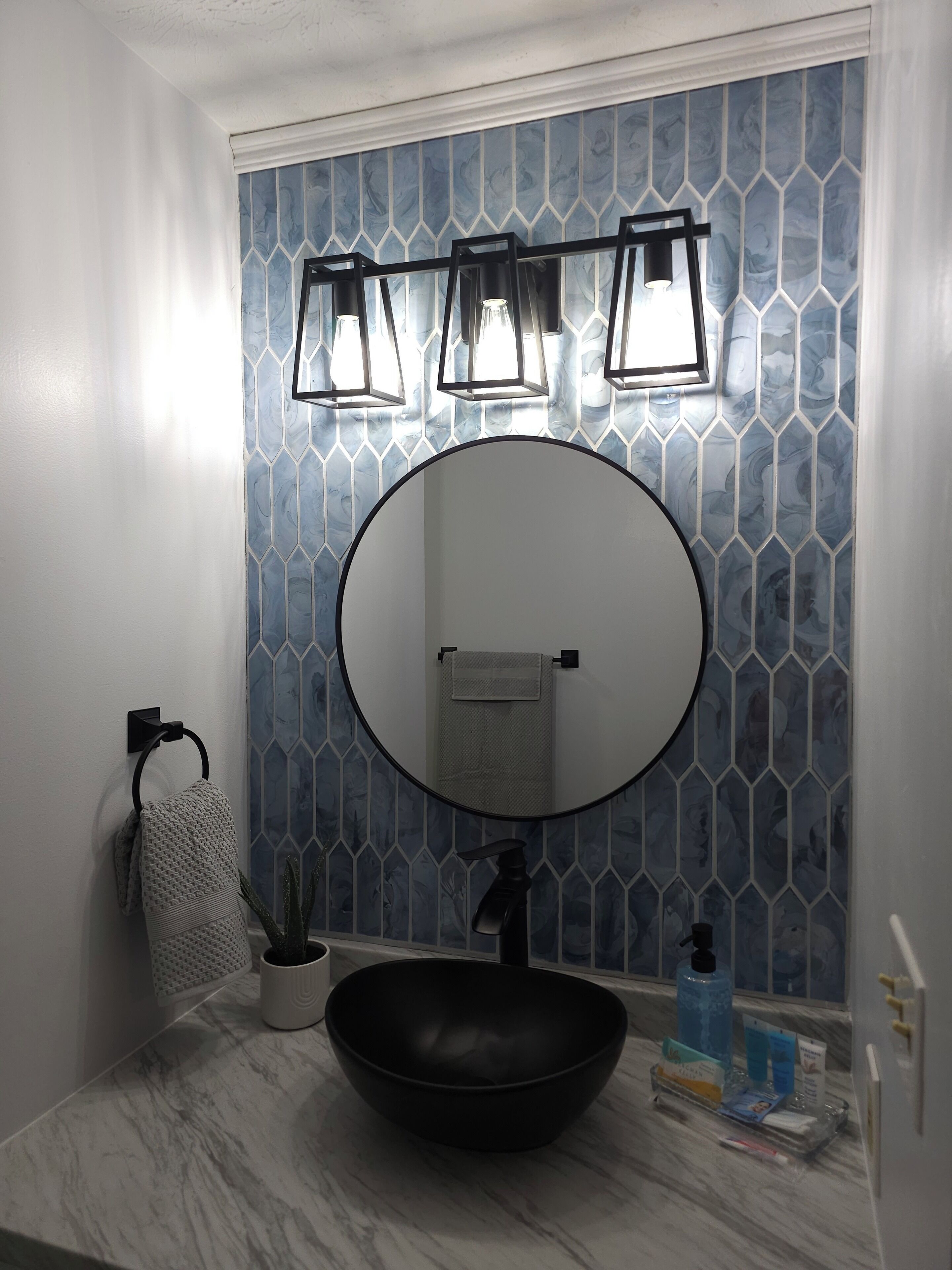 Een badkamer met een ronde spiegel, een zwart wastafelbakje, een handdoekhouder en een wand met een geometrisch tegelpatroon.