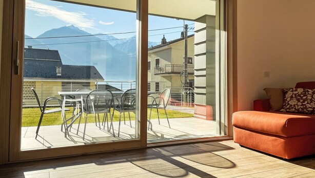 A room with a sliding glass door leading to an outdoor patio with a table and chairs, a red sofa, and a view of mountains.
