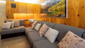 Een kamer met houten lambrisering, een grote bank en een wandtelevisie waarop een mooi landschap te zien is.