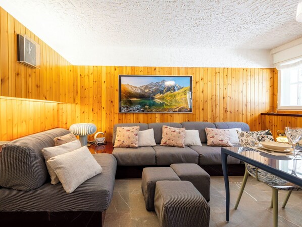 A cozy living room with wooden walls, a large painting of a mountain landscape, a sofa set, and a dining table with a glass.