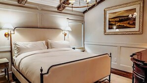 A neatly arranged bedroom with a bed, bedside lamps, a framed landscape painting, and a wooden floor. - The Donna King Suite - Bootleggers Lodge on Lake Nokomis (Tomahawk)