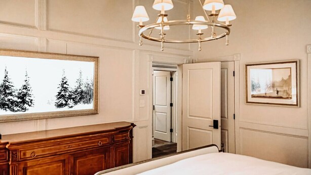 A bedroom with a large bed, a wooden dresser, a chandelier, a window with a view of trees, and a door leading to another room. - The Donna King Suite - Bootleggers Lodge on Lake Nokomis (Tomahawk)