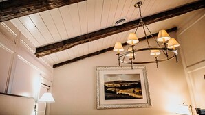 A room with exposed wooden beams, a chandelier with multiple lamps, a framed landscape painting on the wall, and a bed with a bedside lamp. - The Donna King Suite - Bootleggers Lodge on Lake Nokomis (Tomahawk)