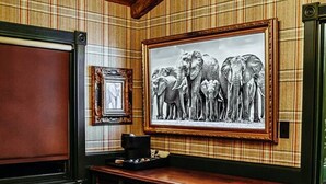 Una habitación con techo de madera, un cuadro grande de un elefante enmarco, un mueble de madera oscura y un banco tapizado en rojo.