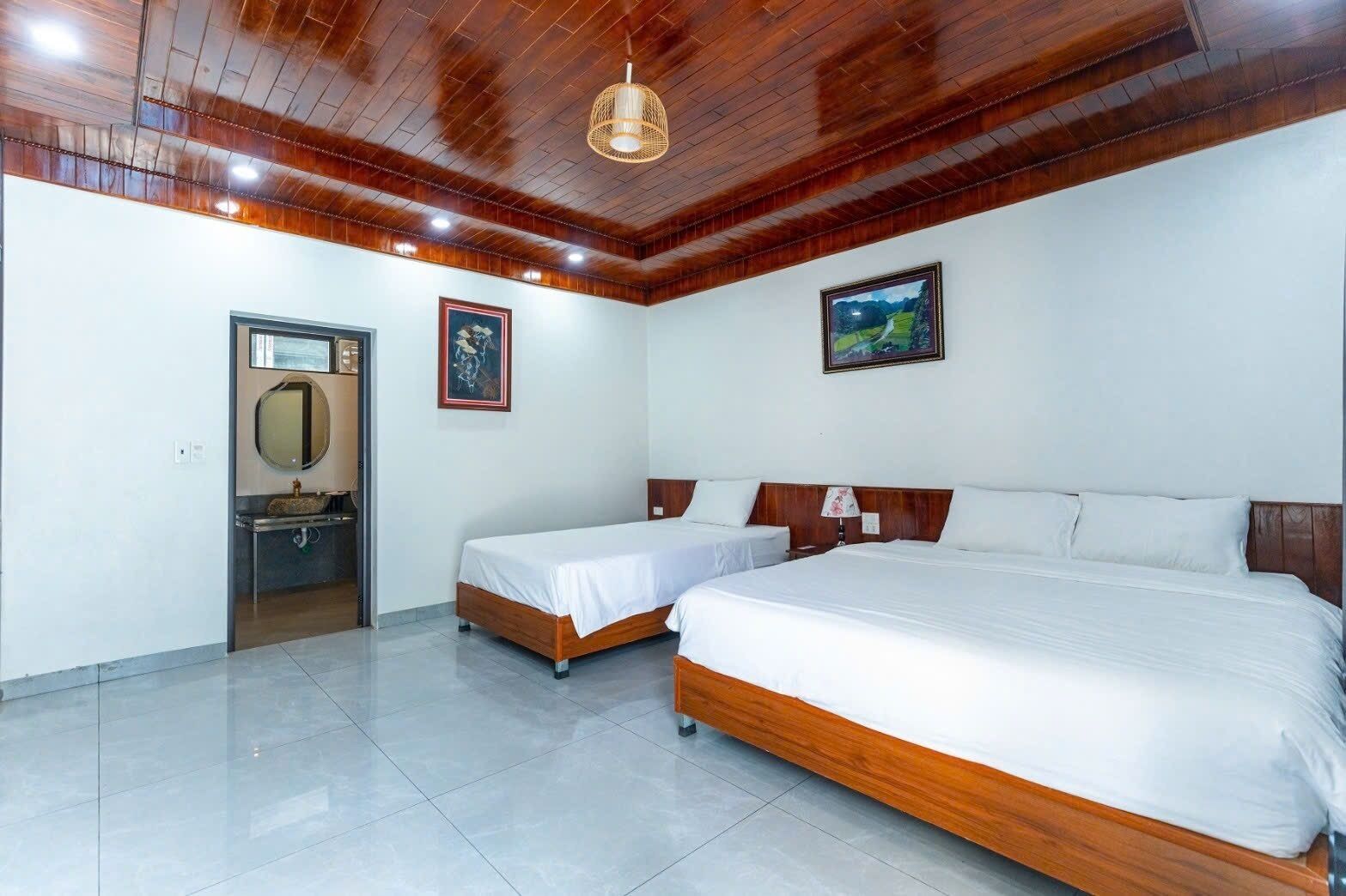 Habitación de hotel con dos camas individuales, techo de madera, baño con espejo y lavamanos, y dos cuadros enmarcados en la pared.