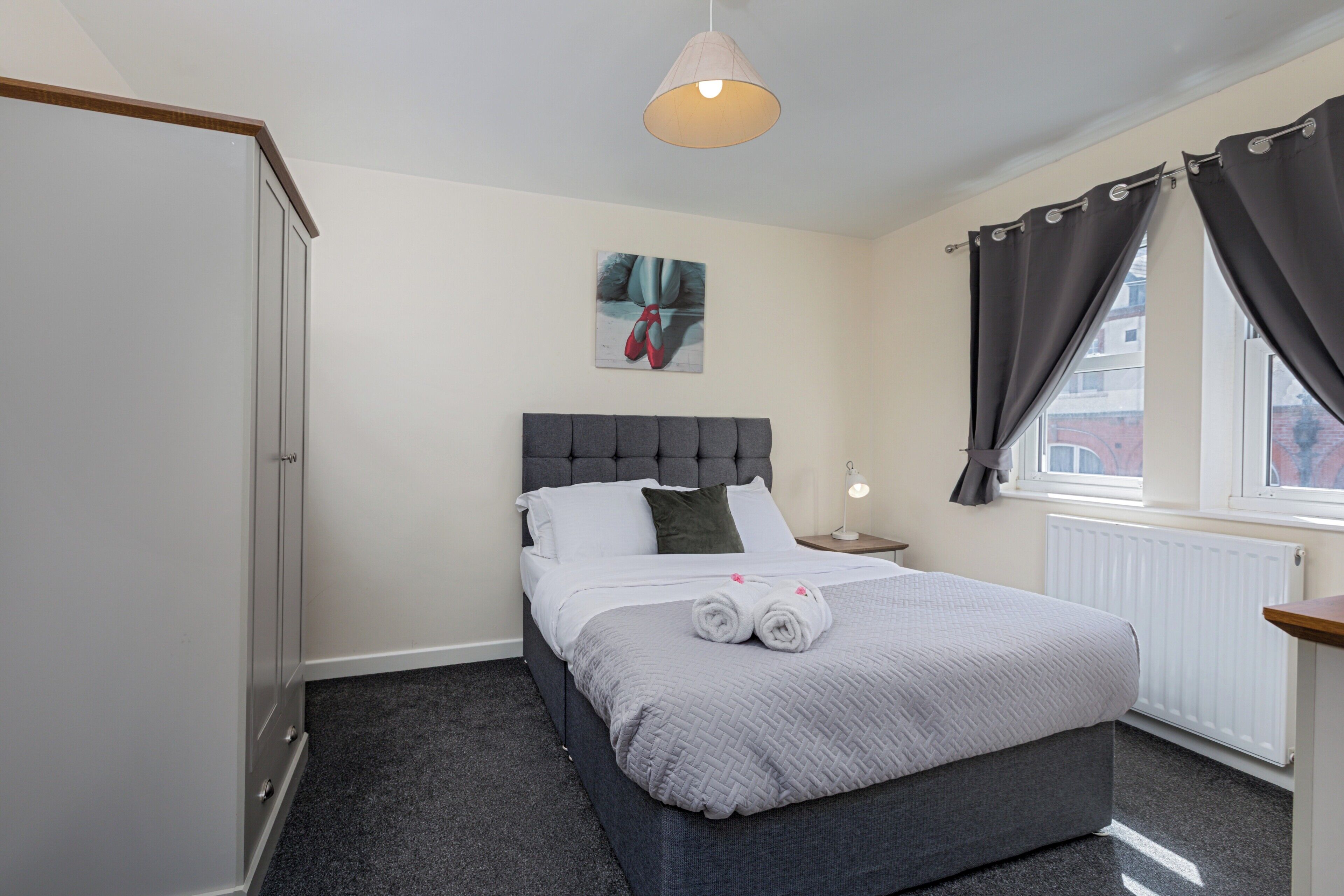 A bedroom with a bed, a nightstand, a lamp, a window with curtains, and a radiator.