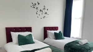 Two twin beds with dark red headboards and green bedding, a wall decal of birds, and a window with blue curtains.