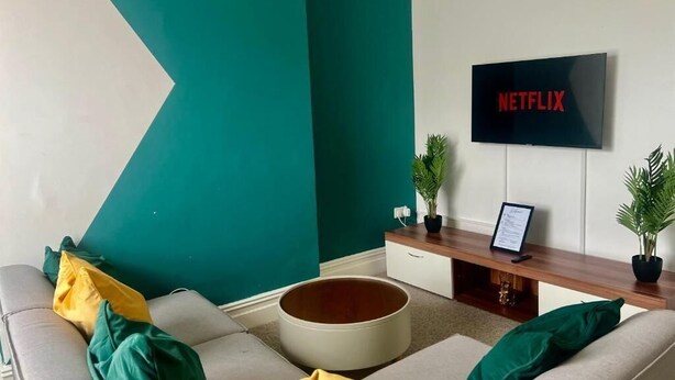 A living room with a teal accent wall, a grey sofa with yellow and green cushions, a round coffee table, and a flat-screen TV mounted on the wall displaying the Netflix logo.