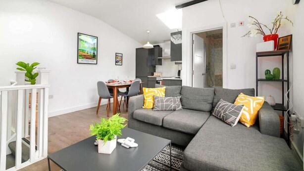 A modern living room with a grey sofa, a dining table with chairs, a black coffee table with a plant, and a kitchen area in the background.