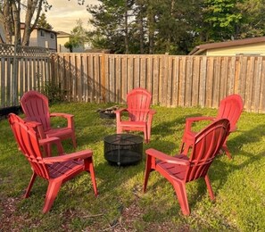 Un giardino recintato da una staccionata di legno, con un braciere e quattro poltrone Adirondack rosse disposte attorno.