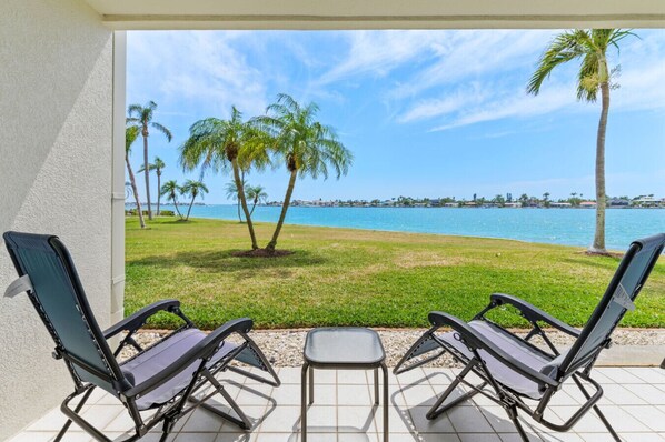 A patio with two lounge chairs and a small table, overlooking a grassy area with palm trees and a view of the water.