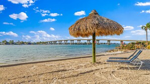A beach with a thatched umbrella, lounge chairs, and a bridge in the background.